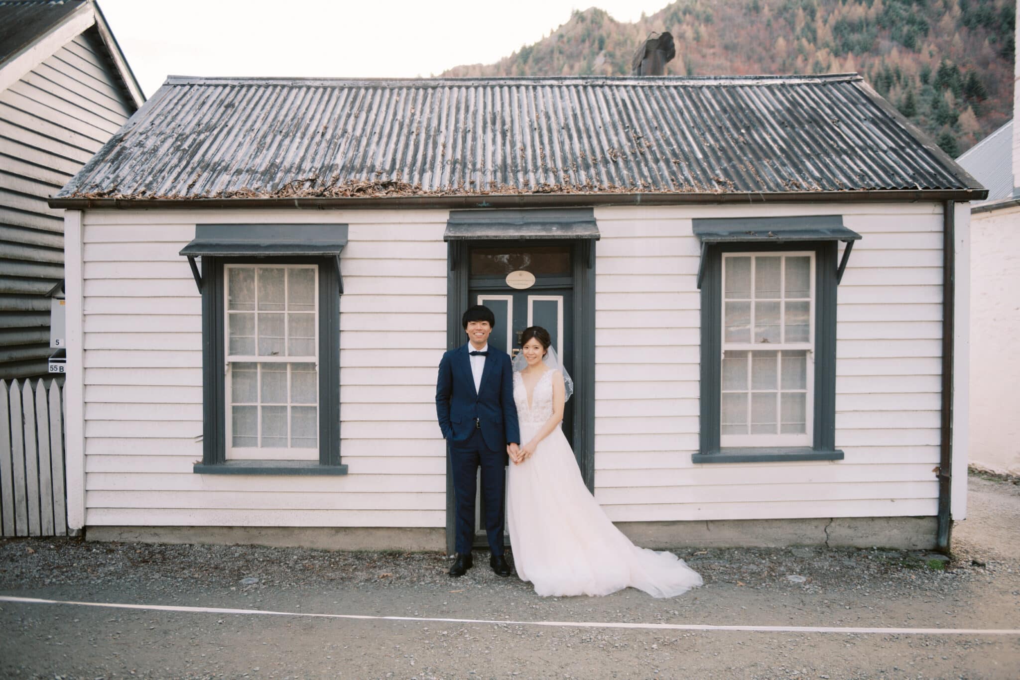 Queenstown Tekapo New Zealand Heli Wedding Elopement Pre-Wedding Shoot Photographer クイーンズタウン　テカポ　ニュージーランド　エロープメント 前撮り　フォトウェディング　結婚式 | A couple dressed in wedding attire standing in front of a quaint, vintage-style house for their Tekapo pre-wedding shoot.